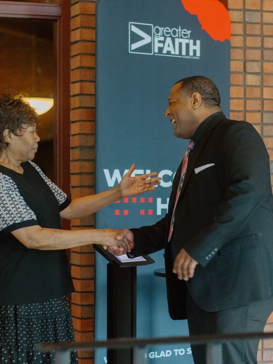 A greeter welcoming a guest with a handshake at the Greater Faith welcome stand